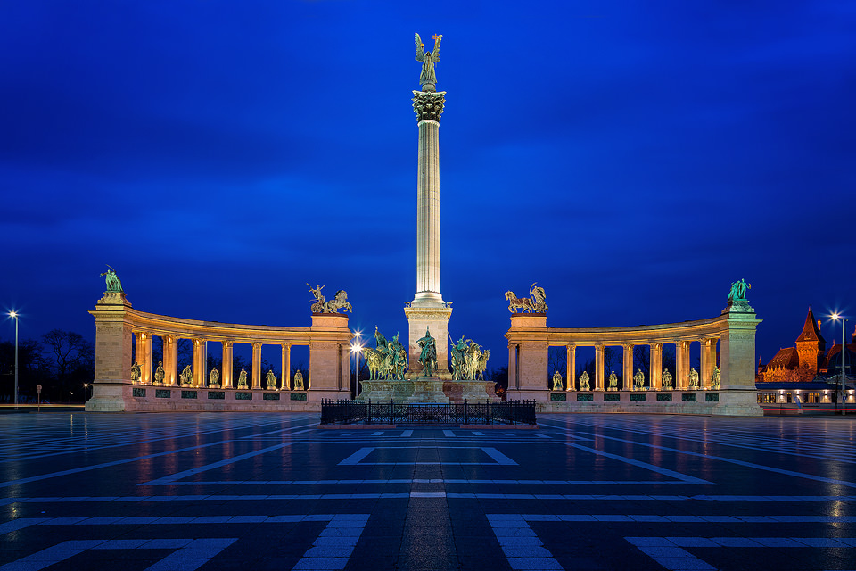 Heroes' Square in Budapest during the blue hour. Heroes’ Square, or Hősök tere in Hungarian, features statues of the fourteen kings of Hungary and a seven metre tall Angel Gabriel on the central pillar. The square looks fantastic lit up at night, making it very popular with tourists, so I had to blend 8 different exposures together to remove the people, creating the apparently deserted scene you see here.